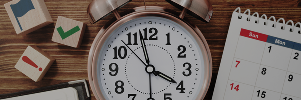 The image appears to focus on a workspace with an alarm clock, some wooden blocks with icons, and a calendar. It seems to convey themes related to time management, planning, and organization.