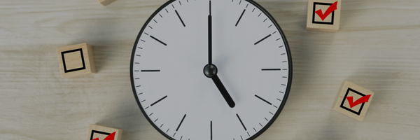 An overhead view of a minimalist wall clock showing 5:00, resting on a light wooden desk. It is surrounded by small wooden blocks—some empty and others stamped with a red checkmark in a square box.